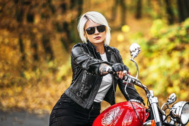 Pretty blonde biker girl in sunglasses with red motorcycle on the road in the forest