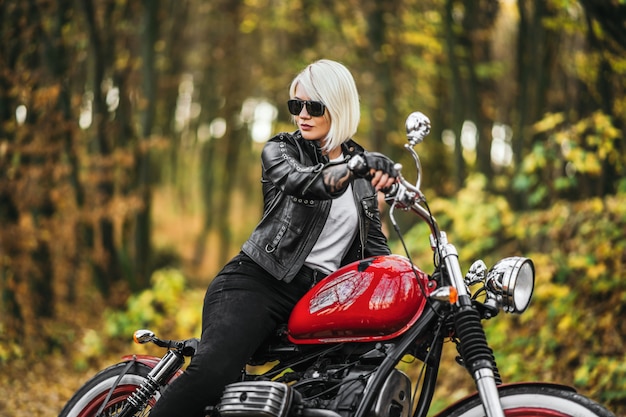 Ragazza bionda graziosa del motociclista in occhiali da sole con il motociclo rosso sulla strada nella foresta