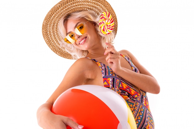 La femmina caucasica bionda graziosa si leva in piedi in costume da bagno con la sfera variopinta, il lolipop e i sorrisi della grande spiaggia di gomma isolati