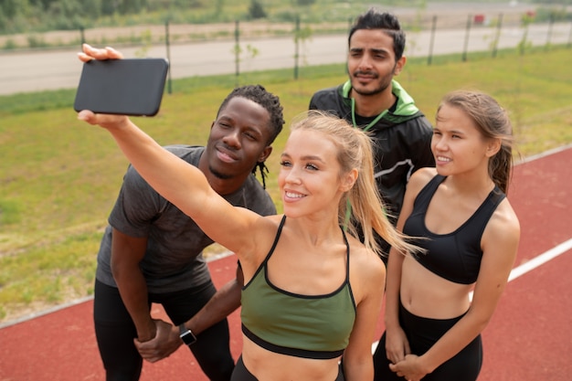 Photo pretty blonde girl in activewear making selfie with her friends standing near by on outdoor stadium