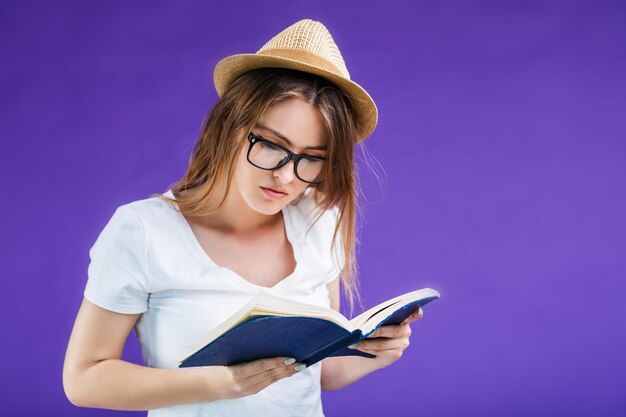 Pretty blonde girl dressed in white tshirt hat and eyeglasses reads book before blue background