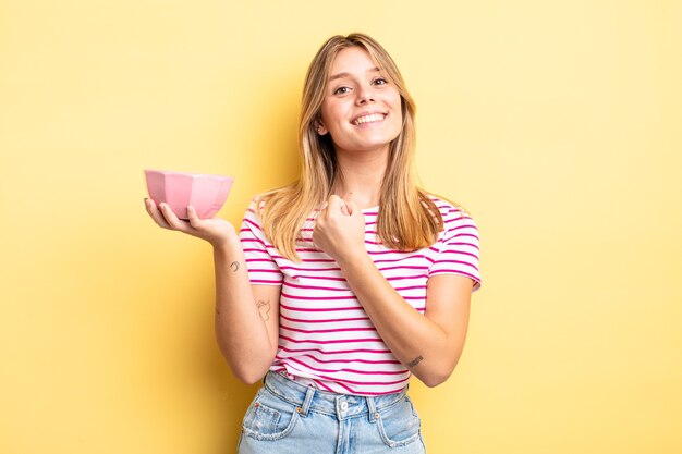 Pretty blonde girl feeling happy and facing a challenge or celebrating. empty bowl concept