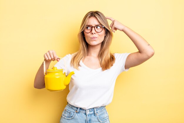 Pretty blonde girl feeling puzzled and confused, scratching head. teapot concept