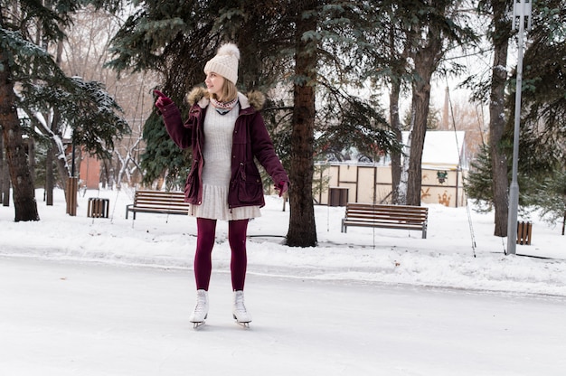 The pretty blonde girl on the figured skates on a opened skating rink in snowy winter park. Winter holidays concept