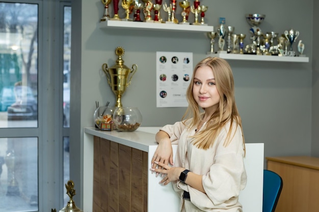 a pretty blonde girl is standing near the reception desk there are cups on the shelves behind