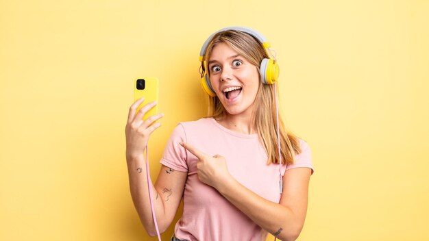 Pretty blonde girl looking excited and surprised pointing to the side. listening music concept