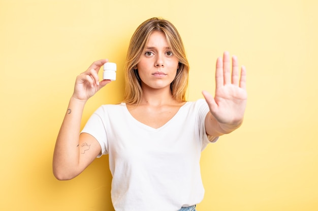 Bella ragazza bionda che sembra seria mostrando palmo aperto che fa gesto di arresto. concetto di bottiglia di pillole