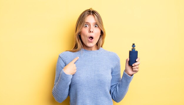 Pretty blonde girl looking shocked and surprised with mouth wide open, pointing to self. smoke vaporizer concept