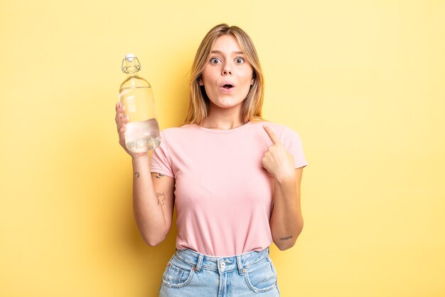 Pretty blonde girl looking shocked and surprised with mouth wide open, pointing to self. water bottle concept