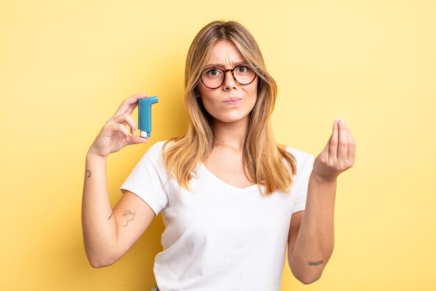 Pretty blonde girl making capice or money gesture, telling you to pay. asthma inhaler concept