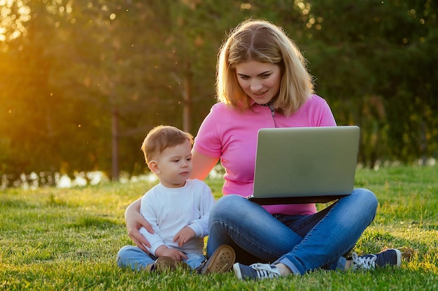 Pretty blonde girl mother and cute son working on a laptop in the park on a background of green grass and trees