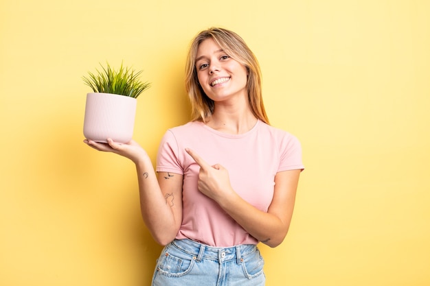 pretty blonde girl smiling cheerfully, feeling happy and pointing to the side. houseplant concept