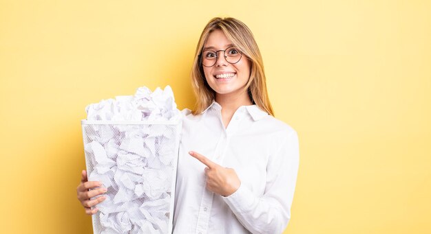 元気に笑って、幸せを感じて、横を指しているきれいなブロンドの女の子。紙球ゴミ箱の概念