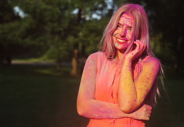 Pretty blonde model wearing orange dress posing covered with a colorful dry paint at the park