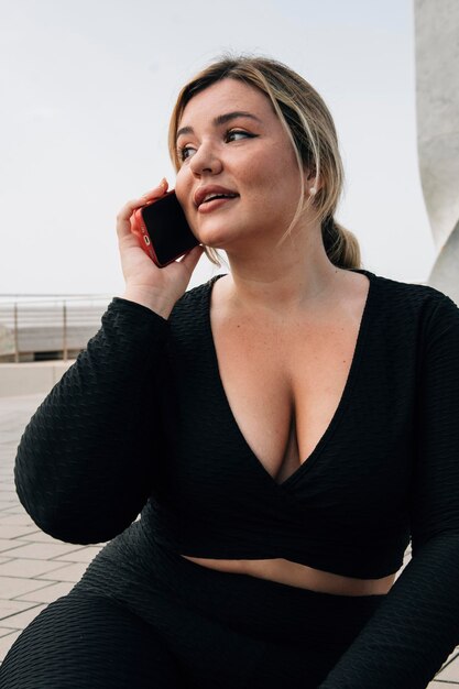 Симпатичная блондинка plus size разговаривает по мобильному телефону перед пляжем, одетая в спортивную одежду