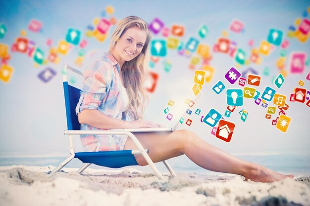 Foto bella bionda seduta sulla spiaggia usando il suo computer portatile con applicazioni informatiche colorate