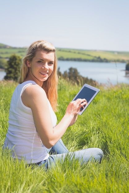 Pretty blonde sitting on grass using her tablet smiling at camera