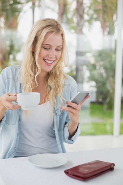 Foto abbastanza biondo che si siede alla tavola che mangia caffè che invia testo