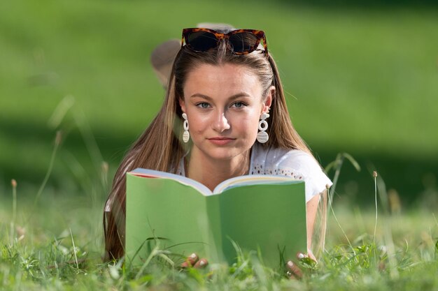 草の上に横たわっている本を学び、読んでいるかなり金髪の学生ポートレートカメラを見て