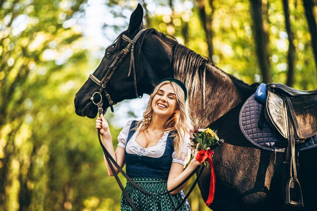 Bionda graziosa in vestito tradizionale che cammina con il grande cavallo nero nella foresta