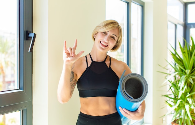 Pretty blonde woman feeling happy, fun, confident, positive and rebellious, making rock or heavy metal sign with hand