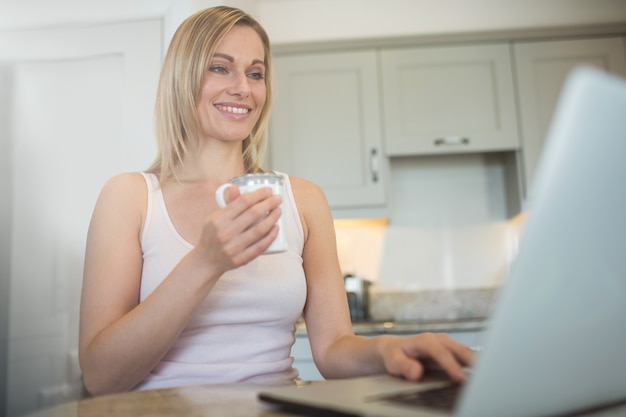 Donna bionda graziosa che mangia caffè e che per mezzo del computer portatile