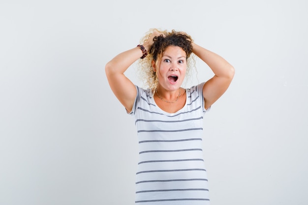 Pretty blonde woman keeping hands on head in striped t-shirt, looking surprised and happy , front view.