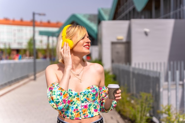Симпатичная блондинка слушает музыку в городе в желтых наушниках и ведет образ жизни модели в городе, где она пьет кофе на вынос