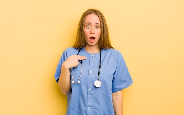 Pretty blonde woman looking shocked and surprised with mouth wide open pointing to self nurse concept