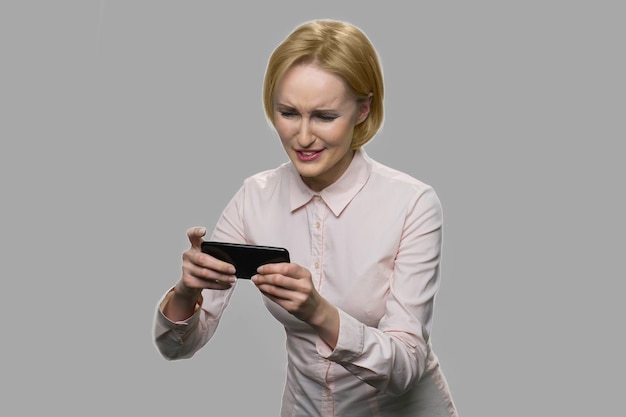 Довольно блондинка играет в онлайн-игру. Смешная бизнес-леди, играя в видеоигры на своем смартфоне на сером фоне.