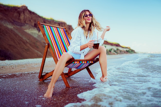 Donna bionda graziosa a forma di un cuore occhiali da sole, camicia bianca e costume da bagno spogliato che si distende su una spiaggia più lettino e beve cocktail. concetto di vacanze estive