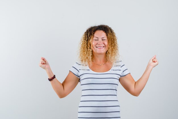 縞模様のTシャツで勝者のジェスチャーを示し、至福に見えるかなり金髪の女性。正面図。