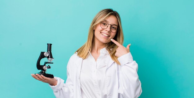 Pretty blonde woman smiling confidently pointing to own broad smile sciencist and microscope concept