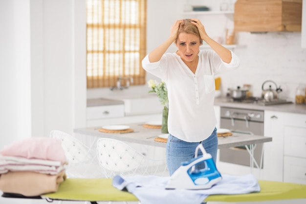 Pretty blonde woman standing near ironing table and looking desperate