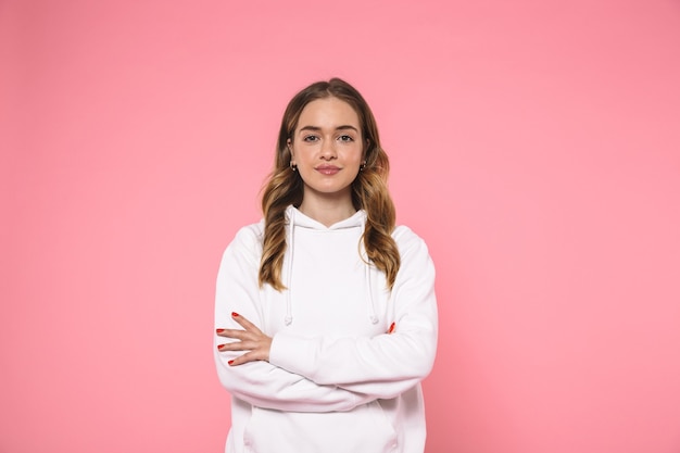 Pretty blonde woman wearing in casual clothes posing with crossed arms and looking at the front over pink wall