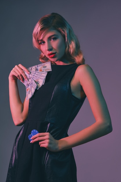 Photo pretty blonde woman with bright makeup in black stylish dress is holding some money and chips while posing against colorful studio background gambling entertainment poker casino closeup