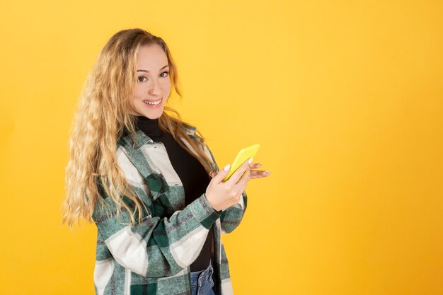 Bella donna bionda con abiti casual, con il suo telefono cellulare sorridente guardando la fotocamera, sfondo giallo