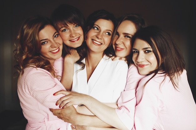 Pretty bride and laughing bridesmaids in pink robes hug each othet tender like sisters 