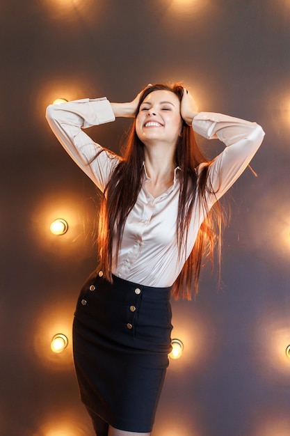 Pretty brunette in blouse and short skirt stands in studio with brick wall and lamps and keeps his hands on his head