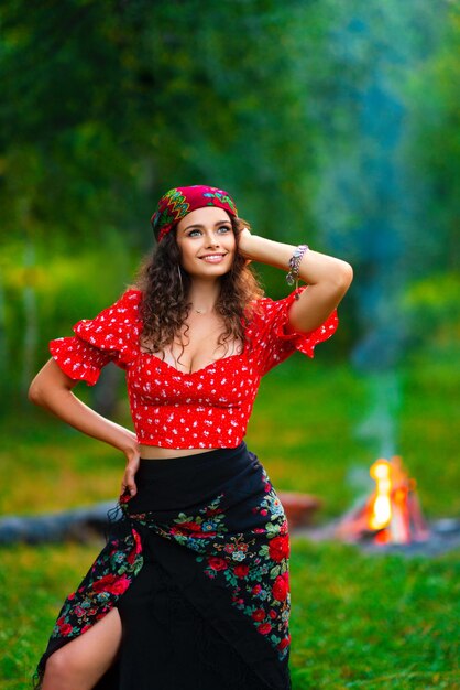 Фото Красивая брюнетка кудрявая девушка в красном цыганском костюме и аксессуарах позирует на природе с костром