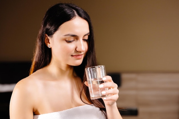 Pretty brunette drinking water standing near bed at home in the bedroom