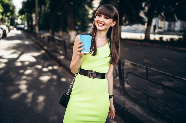커피 한잔과 함께 녹색 드레스에 예쁜 갈색 머리 소녀 거리에서 야외 산책