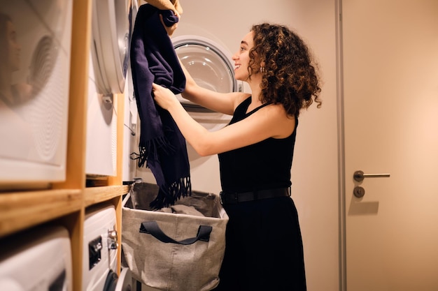 Pretty brunette girl happily loading clothes into washing machine in modern self-service laundry