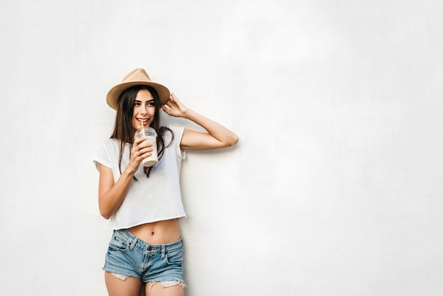 Pretty brunette girl in hat with coffee and white wall behind, copy space.