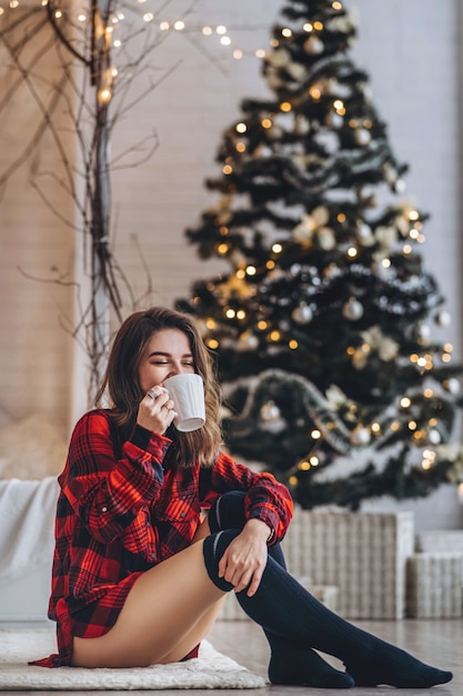 Pretty brunette girl sitting on the floor in red shirt and warm socks with cup of coffee and christmas tree behind