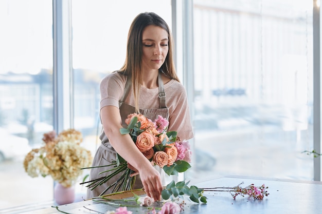 Pretty brunette girl taking one of fresh roses off table while making floral bunches in her studio