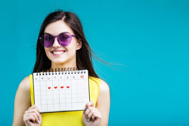 Pretty brunette girl wears yellow shirt and eyeglasses holding vacation calendar on blue background summer time concept