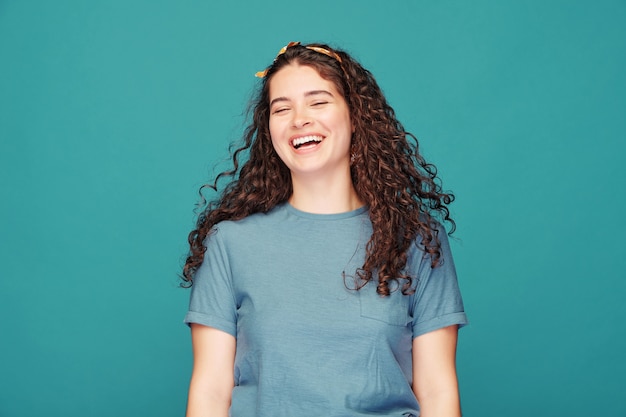 Pretty brunette girl with curly hair laughing loudly on blue, emotion concept