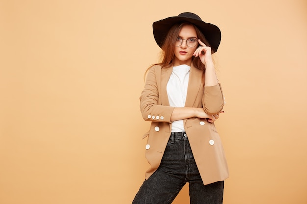 Bella ragazza bruna con i capelli lunghi con gli occhiali e un cappello nero in testa vestita con t-shirt bianca, jeans e giacca beige è in posa sullo sfondo beige in studio.