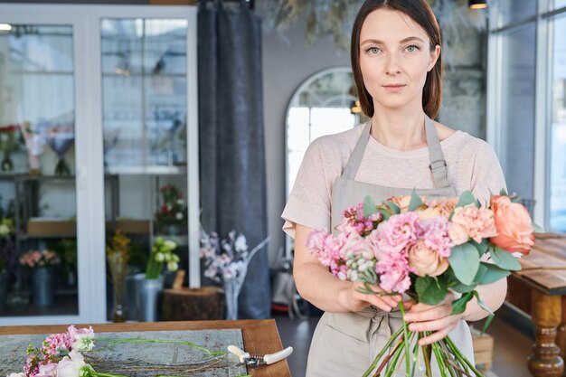 Photo pretty brunette girl in workwear standing in front of camera while preparing fresh flowers for bunches